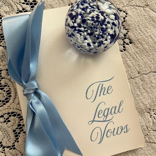 Vows folder
tied with pale blue ribbon. The words The Legal
Vows are on the front. The folder is on an ecru
lace tablecloth. A blue and white paperweight is
also on the table.